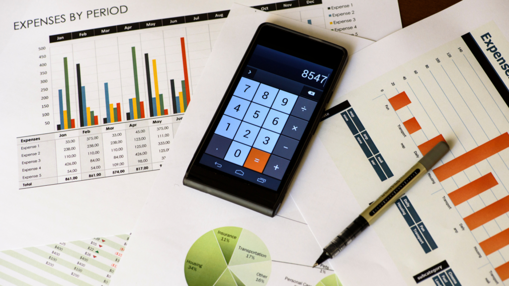 A smartphone with a calculator app open, placed on top of printed sheets featuring various colorful bar graphs and pie charts related to expenses.