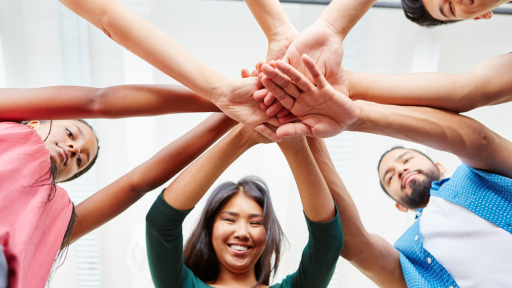 A group of diverse people standing in a circle, with their hands stacked together in the center, smiling and looking down at the camera.