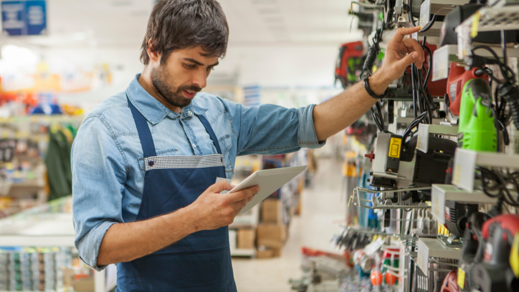 A male store employee in a denim shirt and apron, holding a tablet while examining items on a shelf in a hardware store.