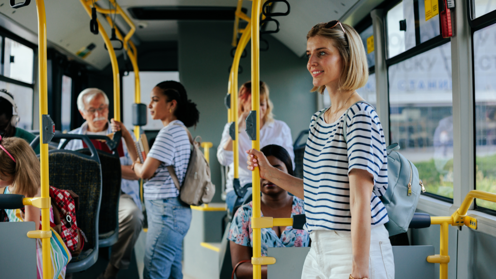 People standing and sitting inside a bus, with a woman in a striped shirt smiling and holding onto a pole.