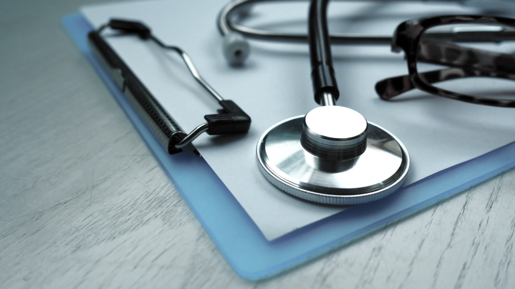 A stethoscope, a pair of glasses, and a clipboard with papers, placed on a light grey surface.