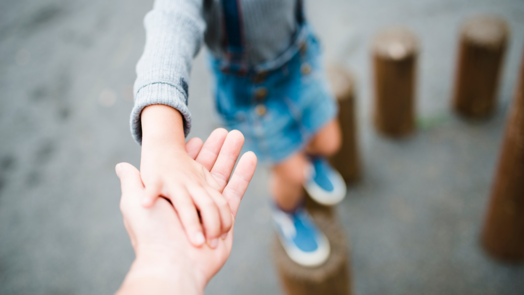 A child holding an adult's hand for support while walking on a path, with the focus on their hands and the child's legs visible in the background.