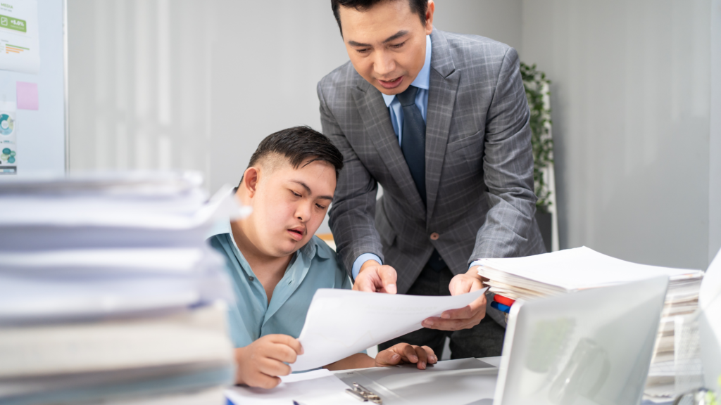 An office setting where a man in a suit is helping a colleague review documents at a desk cluttered with stacks of papers.